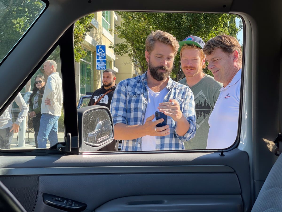 Viewed through the window from within the cab of a vehicle, a man looks at his phone while two others smile and look over his shoulder at the screen.